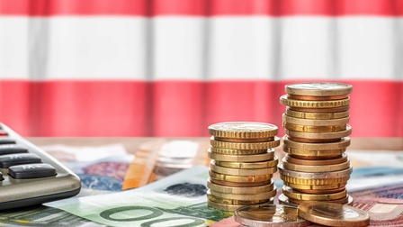 Im Vordergrund sind zwei Münzstapel, unter denen verschiedene Geldscheine liegen. In der linken Bildhälfte ist ein Taschenrechner. Dahinter hängt eine Flagge in den Farben rot-weiß-rot