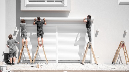 Drei Personen auf Holzstehleitern halten Markisenpaneelzur Befestigung über bodentiefe Fenster mit heruntergelassenen Außenjalousien an weiße Hausfassade, weitere Person am Boden stehend blickt zu ihnen hinauf