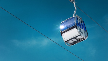 Weiße Seilbahngondel in deren Fenster sich Sonne spiegelt, an Seil hängend unter blauem Himmel