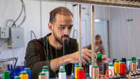 Eine Person steht vor vielen, bunten, stehenden Garnrollen, unter denen ein Tisch ist. Von jeder Rolle geht ein Faden nach oben zu einer Halterung. Die Person hält einen Garn in den Händen und blickt darauf