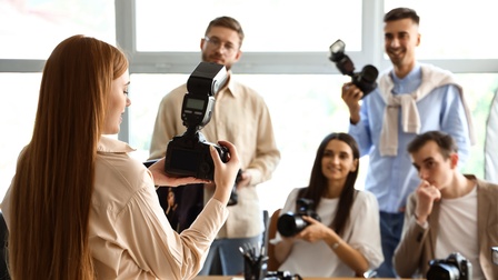 Rückenansicht einer Person, die einen Fotoapparat in beiden Händen hält. Der Person gegenüber sind vier weitere Personen, die ebenfalls jeweils einen Fotoapparat halten.