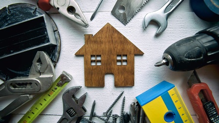 Verschiedene Werkzeuge liegen auf einem Holztisch in einem runden Kreis um ein kleines Modellhaus.