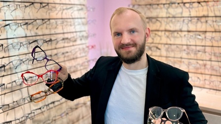 Raphael Titz in seinem Laden vor Brillen-Hintergrund, er hält eine Brille ins Bild