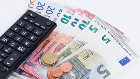 Eine Fernbedienung liegt auf nebeneinander aufgelegten Geldscheinen am Tisch, daneben sind noch ein paar Münzen gestapelt.