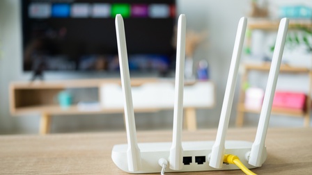Auf einem Holztisch steht ein WLAN-Router, aus dem zwei Kabel kommen. Im Hintergrund ist ein großer Bildschirm