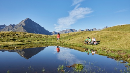 Zwei Personen in Wiese an Bergsee sitzend, eine weitere anbei stehend, spiegeln sich in Gewässer, im Hintergrund Bergkette und blauer Himmel