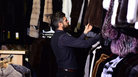 Eine Person in einem dunklen Hemd, mit dunklen Haaren und Bart steht in einem Geschäft an Kleiderstangen und hält mit der linken Hand eine Pelzjacke. Daneben hängen weitere Produkte aus Pelz