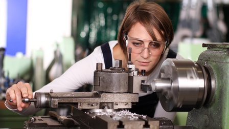 Person mit Schutzbrillen bedient Drehmaschine, in der Metallstück geschliffen wird und blickt auf Werkstück