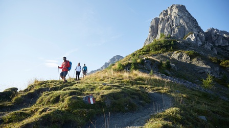 Drei Personen in Wanderkleidung, zwei davon mit Wanderstöcken ausgerüstet, marschieren Berg herunter