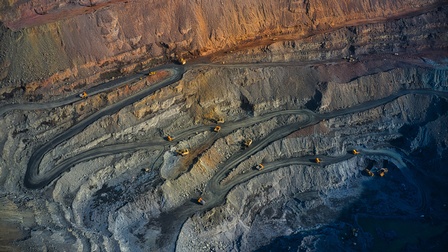 Aufnahme eines Bergwerks aus der Luft mit verschlungenen Wegen, die vom Rand des Werks nach unten führen. Auf den Wegen fahren mehrere Fahrzeuge