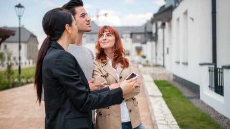 Seitliche Aufnahme dreier Personen, die vor einem Haus stehen. Die Person links hält ein Tablet. Im Hintergrund sind weitere Häuser und ein gepflasteter Weg