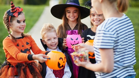 Vier Kinder in unterschiedlichen Kostümen stehen nebeneinander auf einem asphaltierten Weg. Drei Kinder halten einen Plastikkürbis vor ihre Oberkörper. Neben ihnen steht eine erwachsene Person, die eine kleine Plastikschale mit Süßigkeiten hält
