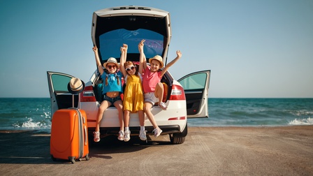 Drei Kinder mit Hüten und Sonnenbrillen sitzen im geöffneten Kofferraum eines am Strand vor Meer parkenden Autos und strecken jubelnd die Arme in die Luft, Türen des Wagens sind geöffnet, vor dem Auto oranger Rollkoffer mit Strohhut abgestellt