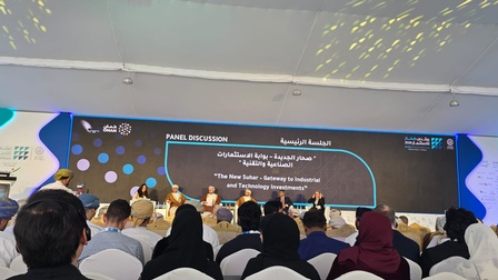 Rückenansicht eines Publikums. Dem Publikum gegenüber sitzen mehrere Personen auf einem Podium auf Sesseln nebeneinander. Hinter ihnen ist ein großer Bildschirm, auf dem Das neue Suhar - das Tor zu industriellen und technologischen Investments