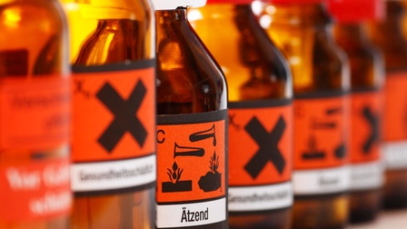 Mehrere braune Flaschen nebeneinandergereiht mit orangen Etiketten und schwarzem X, im Fokus eine Flasche mit der Aufschrift ätzend und Symbolen