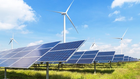 Mehrere Reihen von Solarpanelen auf Metallgerüsten auf einem Feld, dahinter Windräder unter blauem Himmel mit vereinzelten Wolken