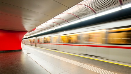 (KI-generierter Alttext) U-Bahn fährt mit Bewegungsunschärfe an einem leeren Bahnsteig in einem modernen Bahnhof mit roter Wand und Deckenbeleuchtung vorbei.