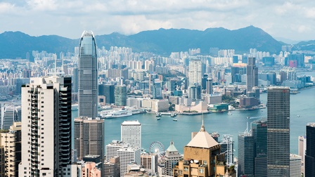 Stadtansicht von Hongkong: Blick von oben auf die am Wasser gelegene Stadt mit etlichen Wolkenkratzern unter bewölktem Himmel und Bergen im Hinterland. 