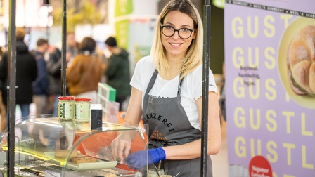 Porträt einer Person mit blonden schulterlangen Haaren, Brillen und Arbeitsschürze steht bei einem Stand und bereitet Speisen zu, während sie in die Kamera blickt