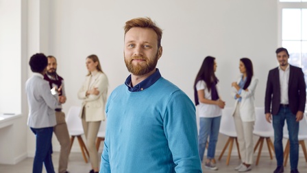 Person mit rötlichem Haar und Bart sowie blauem Pullover blickt freudig in die Kamera, im Hintergrund stehen weitere Personen und unterhalten sich in der Unschärfe