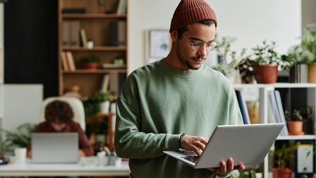 Halbporträt einer Person mit Brille und Mütze, die mit der linken Hand einen aufgeklappten Laptop hält, die rechte Hand auf dessen Tastatur hat und auf die Tastatur blickt. Hinter ihr sitzt eine Person an einem aufgeklappten Laptop auf einem Tisch