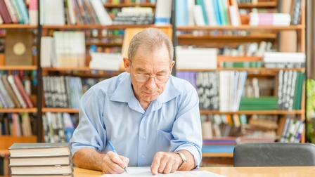 Person mit Brillen sitzt in Bibliothek vor aufgeschlagenem Buch, hält Finger an Passage des geöffneten Buches und nimmt mit anderer Hand Notizen vor