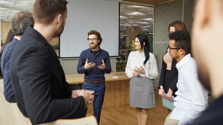 Personen stehen im Kreis und unterhalten sich in einem Besprechungsraum mit Leinwand