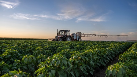 Ein Traktor fährt über ein Feld mit Pflanzen und verteilt über dieses mit einer Vorrichtung Pestizide