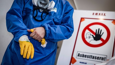 Mann säubert Gebäude von Asbest im blauen Schutzanzug mit gelben Handschuhen und einer Schutzmaske.  