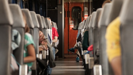 Innenaufnahme eines Zugwagons: Links und Rechts vom Gang graue Sitzplätze in zahlreichen Reihen hintereinander, Menschen im Ausschnitt darauf sitzend erkennbar, im Hintergrund rote Wagontüre
