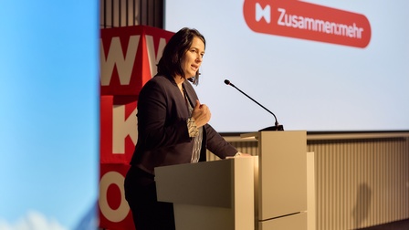Eine Person steht hinter einem Rednerpult und spricht in ein Mikrofon. Die Person trägt ein dunkles Sakko und ein gemustertes Oberteil und hält eine Hand in Brusthöhe, als würde sie etwas betonen. Im Hintergrund ist eine große Projektionsfläche mit dem Wort „Zusammenmehr“ und einem roten Logo zu sehen. Links im Bild steht ein roter Würfel mit weißen Buchstaben. Der Raum wirkt wie ein moderner Veranstaltungssaal mit Bühnenbeleuchtung.