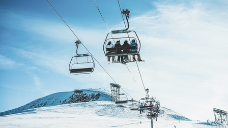 Auf einem Sessellift sitzen auf mehreren Sesseln sitzen Personen in Skianzügen und mit Ski an den Füßen. Unter ihnen sind schneebedeckte Pisten, auf denen vereinzelte Personen sind. Im Hintergrund sieht man schneebedeckte Berge und blauen Himmel.