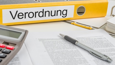 Nahaufnahme eines gelben Ordners, der auf einem weißen Tisch liegt. Auf der Seite des Ordners steht Verordnung. Vor dem Ordner sind bedruckte Zettel, ein Stift und ein Taschenrechner