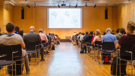 In einem großen Raum sind mehrere Personen, die in vielen Sesselreihen neben- und hintereinander sitzen. Ihnen gegenüber ist eine Person hinter einem Rednerpult. Neben ihr ist eine Leinwand.