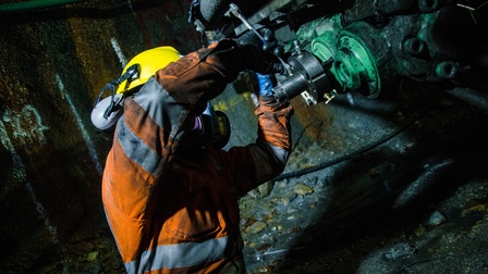 Seitliche Aufnahme einer Person mit Helm, Gehörschutz und reflektierender Kleidung, die in einem Bergwerk in einem Tunnel hockt und einen Schraubschlüssel an eine Maschine hält