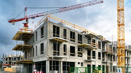 Eine Baustelle eines größeren Gebäudes mit zwei Stockwerken neben dem Kräne stehen. Um die Baustelle ist ein Gitterzaun. Im Hintergrund ist eine weitere Baustelle eines Gebäudes