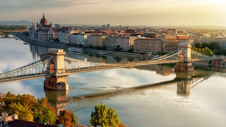 Eine Brücke führt über ein Gewässer. An der rechten Uferseite sind mehrere historische Gebäude. Die linke Uferseite ist nur teilweise im Bild. Direkt am Ufer sind grüne Bäume. Dahinter sind Häuser