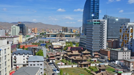 Stadtansicht von Ulan Bator: Blick auf den Choijin Lama Tempel mit Kloster in der Mongolei. Daneben moderne Wolkenkratzer, Berge im Hintergrund unter blauem Himmel.