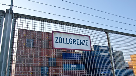 Auf einem Zaun ist ein Schild befestigt, auf dem Zollgrenze steht. Über dem Zaun ist ein Maschendrahtzaun. Dahinter stehen mehrere gestapelte Container