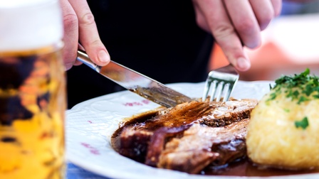Nahaufnahme eines Stücks Fleisch in brauner Sauce. Neben dem Fleisch liegt ein Knödel bestreut mit Petersilie. Eine Person schneidet gerade mit einem Messer in das Fleisch und hält in der linken Hand eine Gabel, womit das Fleisch fixiert wird
