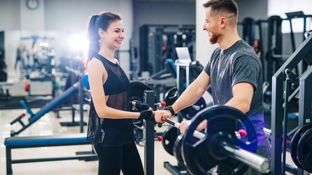 Zwei lachende Personen stehen in Fitnessstudio an Gewichtstemmgerät