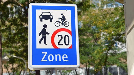 Blauweißes Verkehrsschild mit weißer Umrandung für Begegnungszonen: Schwarze Symbole einer gehenden und einer Person auf einem Fahrrad sowie eines Autos , daneben im rechten unteren Eck die Zahl zwanzig in rotem Kreis, darunter Schriftzug Zone.