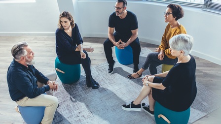 Mehrere Personen sitzen im Kreis auf blauen Hockern und unterhalten sich