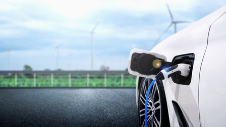 Ein Elektroauto ist an eine Ladesäule geschlossen und wird mithilfe eines Ladekabels aufgeladen, im Hintergrund verschwommen eine grüne Wiese und Windkrafträder.