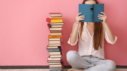 Person sitzt vor pinker Wand  im Schneidersitz am Boden und hält blaues Buch vor Gesicht, daneben Bücherstapel