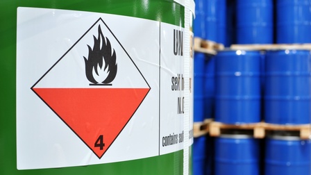 Detailansicht einer grünen Tonne mit Gefahrenaufkleber brennbar, im Hintergrund verschwommen weitere blaue Tonnen aufeinander gestapelt