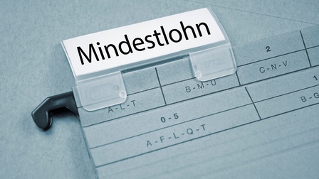 Nahaufnahme eines Ordners zum Aufhängen, worauf ein Plastikschieber befestigt ist. Darin befindet sich ein Stück Papier, bedruckt mit dem Wort Mindestlohn