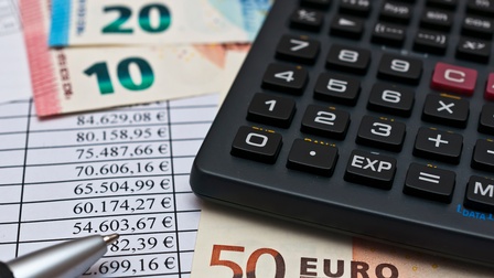 Detailansicht eines Taschenrechners, Euro-Banknoten, Stiftspitze und Unterlagen mit Zahlentabellen