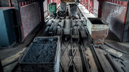 In einer Mine verlaufen zwei Gleise nebeneinander. Auf den Gleisen stehen fünf Karren leicht versetzt. Ein Karren ist mit Kohlestücken gefüllt