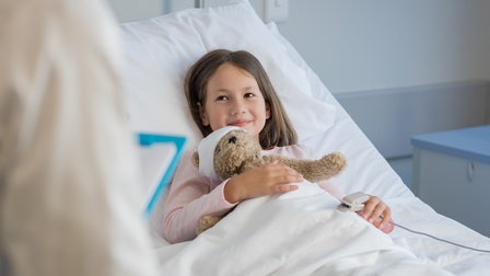 Ein Kind liegt in einem Bett mit weißer Bettwäsche. Auf ihrem Oberkörper liegt ein Teddybär mit verbundenem Kopf. Am linken Finger des Kindes ist ein hellgraues Objekt